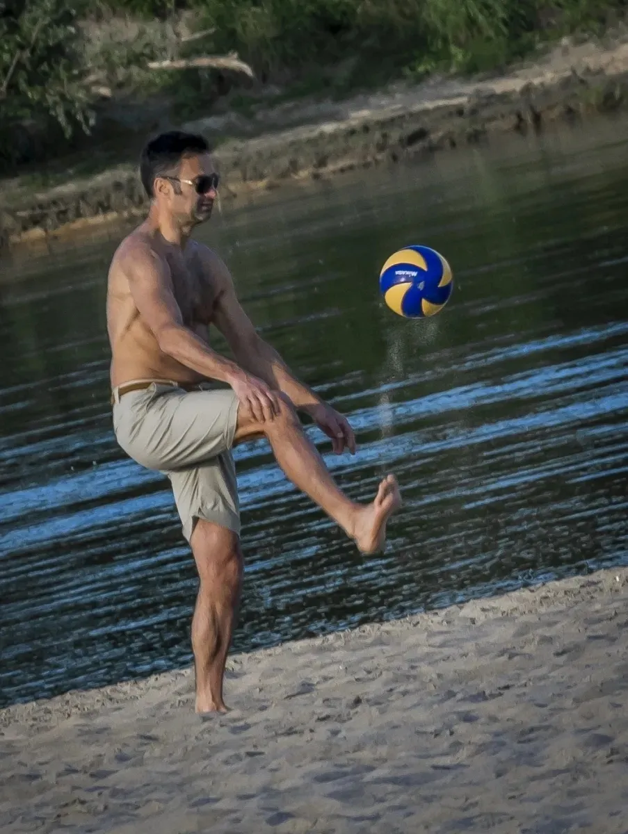 Shirtless man kicking a volleyball on the river beach